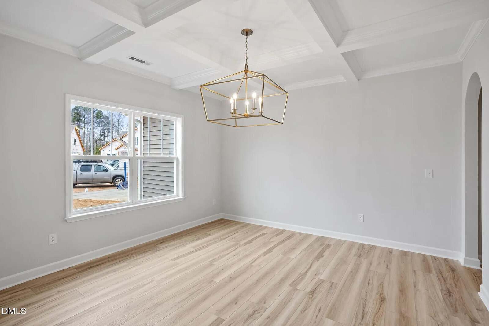 A bright, empty room with light wood flooring, white walls, a large window, and a modern gold geometric chandelier. The ceiling has a coffered design, and an arched doorway is visible on the right.