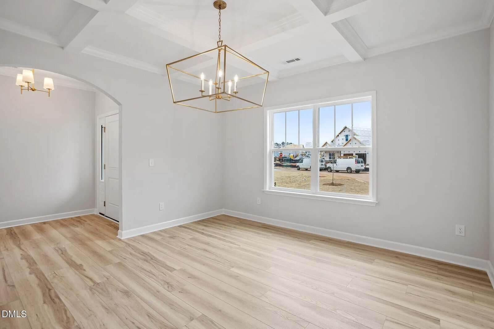 A bright, empty room with light wood floors, white walls, a coffered ceiling, a large window, and a modern gold chandelier. A doorway leads to another room with similar flooring and a hanging light fixture.