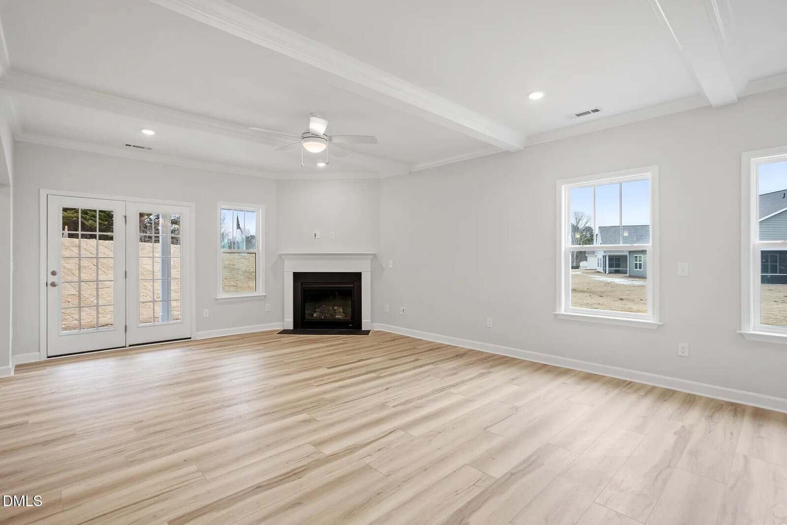 Spacious, empty living room with light wood flooring, white walls, a ceiling fan, a black fireplace, multiple windows, and double glass doors opening to the outside.