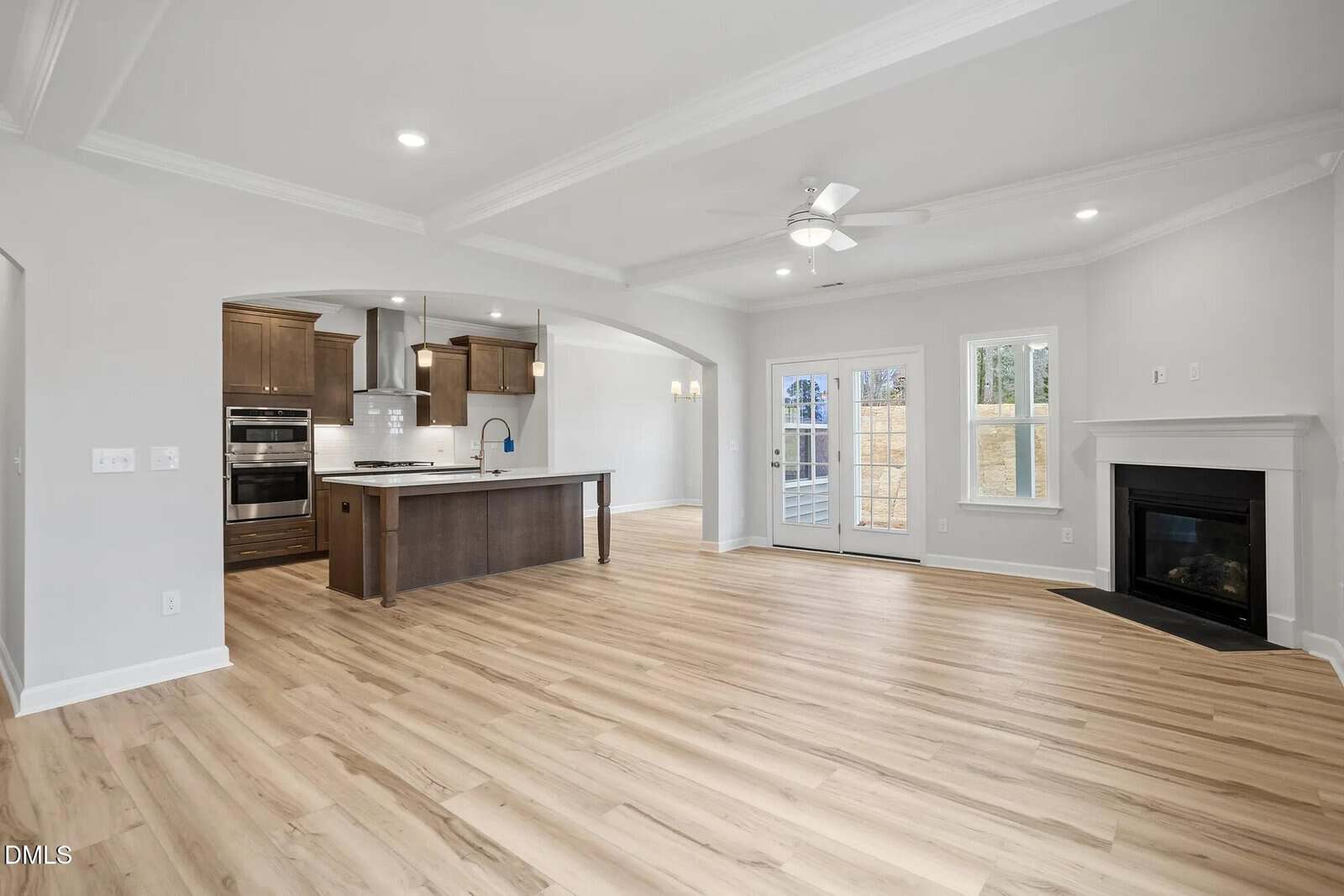 Open-concept living space featuring light wood flooring, a modern kitchen with dark wood cabinets and an island, a ceiling fan, French doors, and a corner fireplace under recessed lighting.