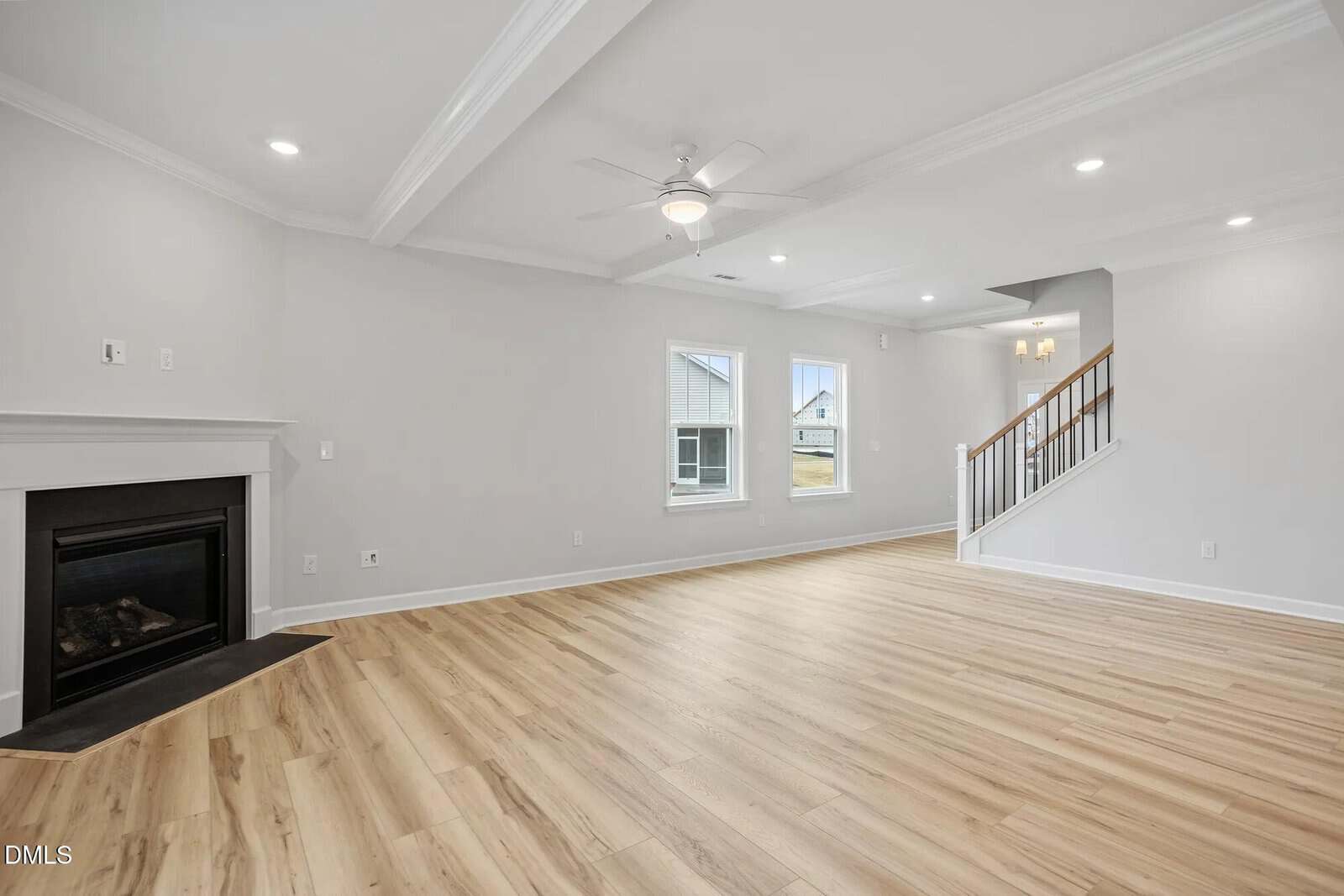 Spacious living room with light wood flooring, white walls, a black fireplace on the left, two windows, a ceiling fan with lights, and a staircase with wooden handrails in the background.