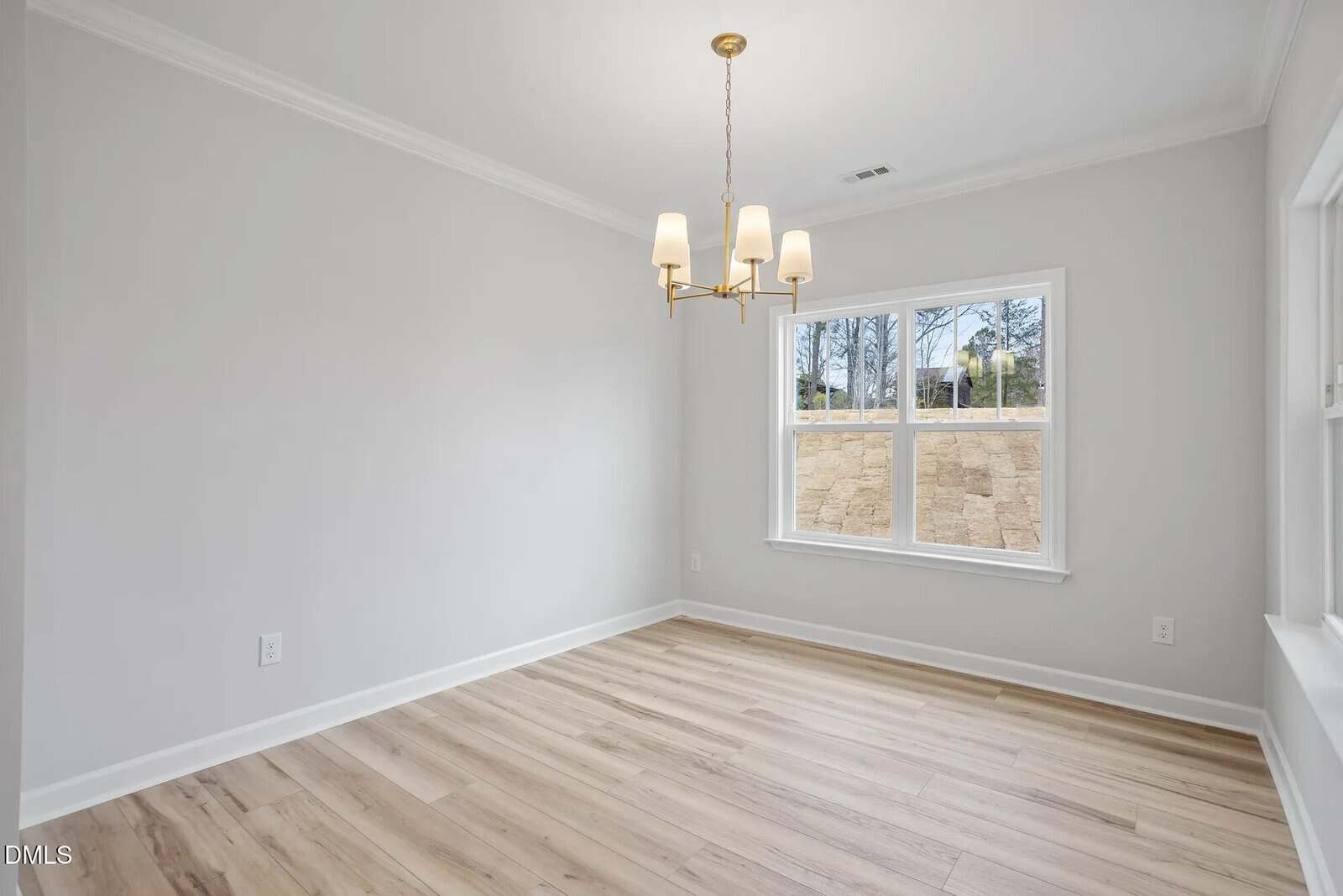 A bright, empty room with light wood flooring, white walls, a white ceiling with crown molding, a modern gold chandelier, and a window showing an outdoor stone retaining wall and trees.