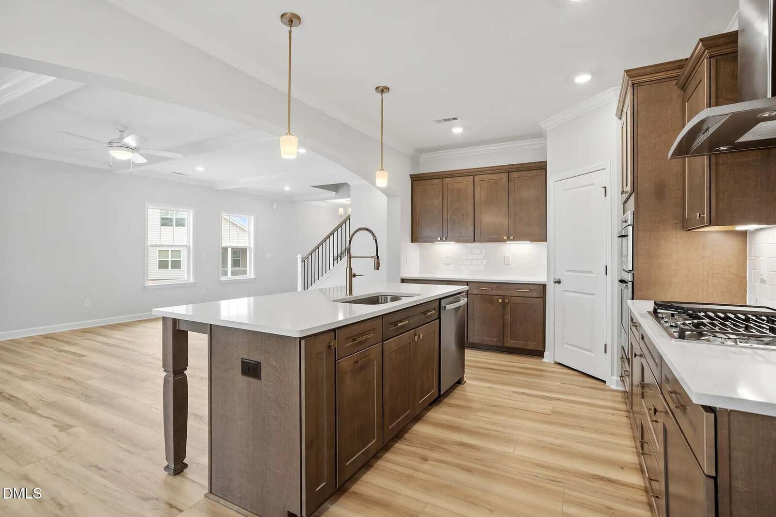 A modern kitchen with wood cabinets, a large island with sink, white countertops, tile backsplash, gas stove, built-in oven, and pendant lights. The kitchen opens to a bright living area with wood floors.