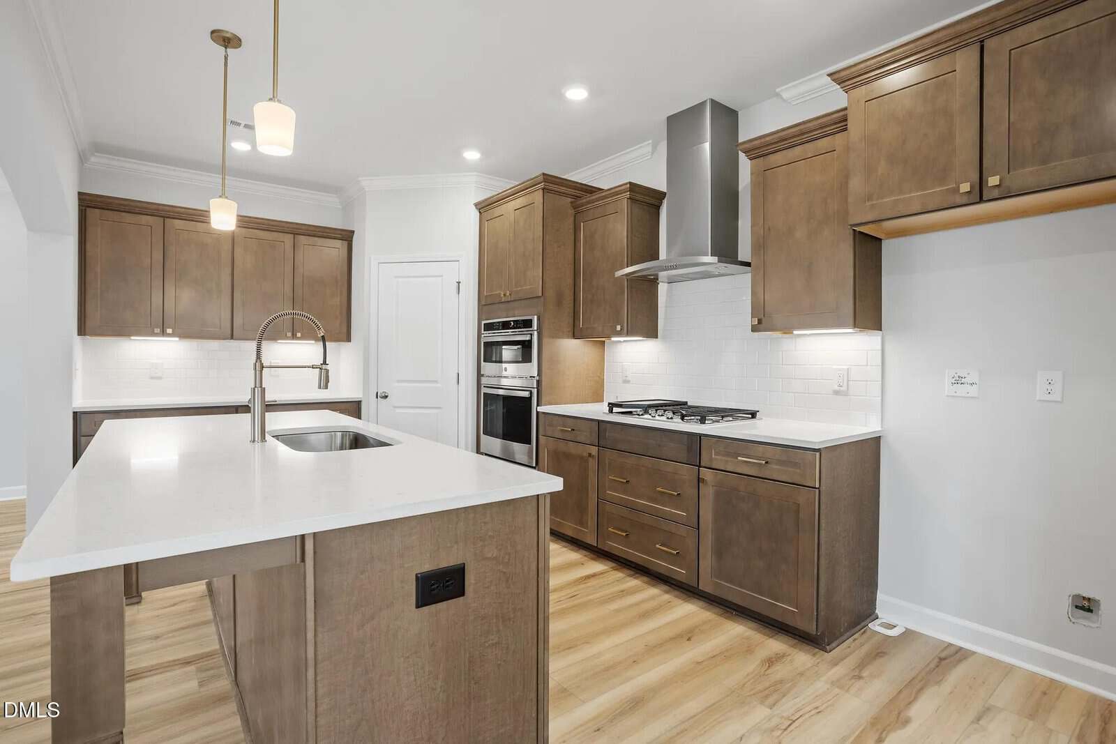 Modern kitchen with brown cabinets, white countertops, an island with a sink, built-in oven, gas cooktop, stainless steel range hood, white subway tile backsplash, and light wood flooring.
