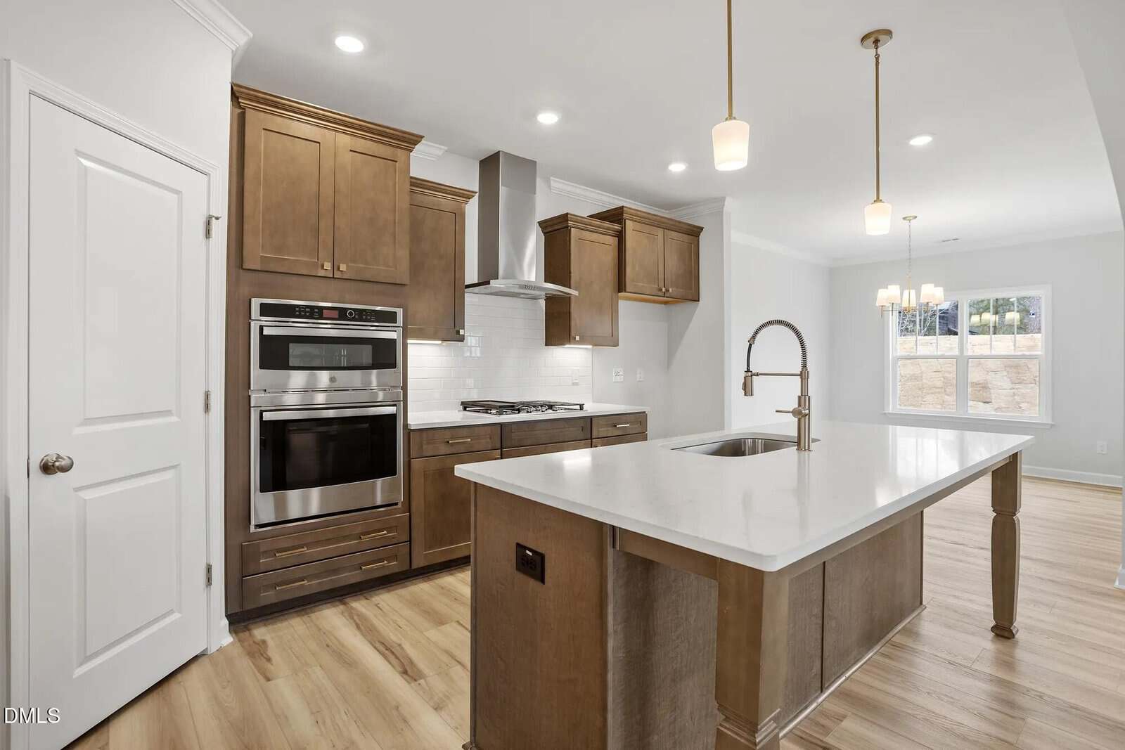 Modern kitchen with wood cabinets, stainless steel appliances, white countertops, an island with a sink and faucet, pendant lights, light wood floors, and a large window providing natural light.
