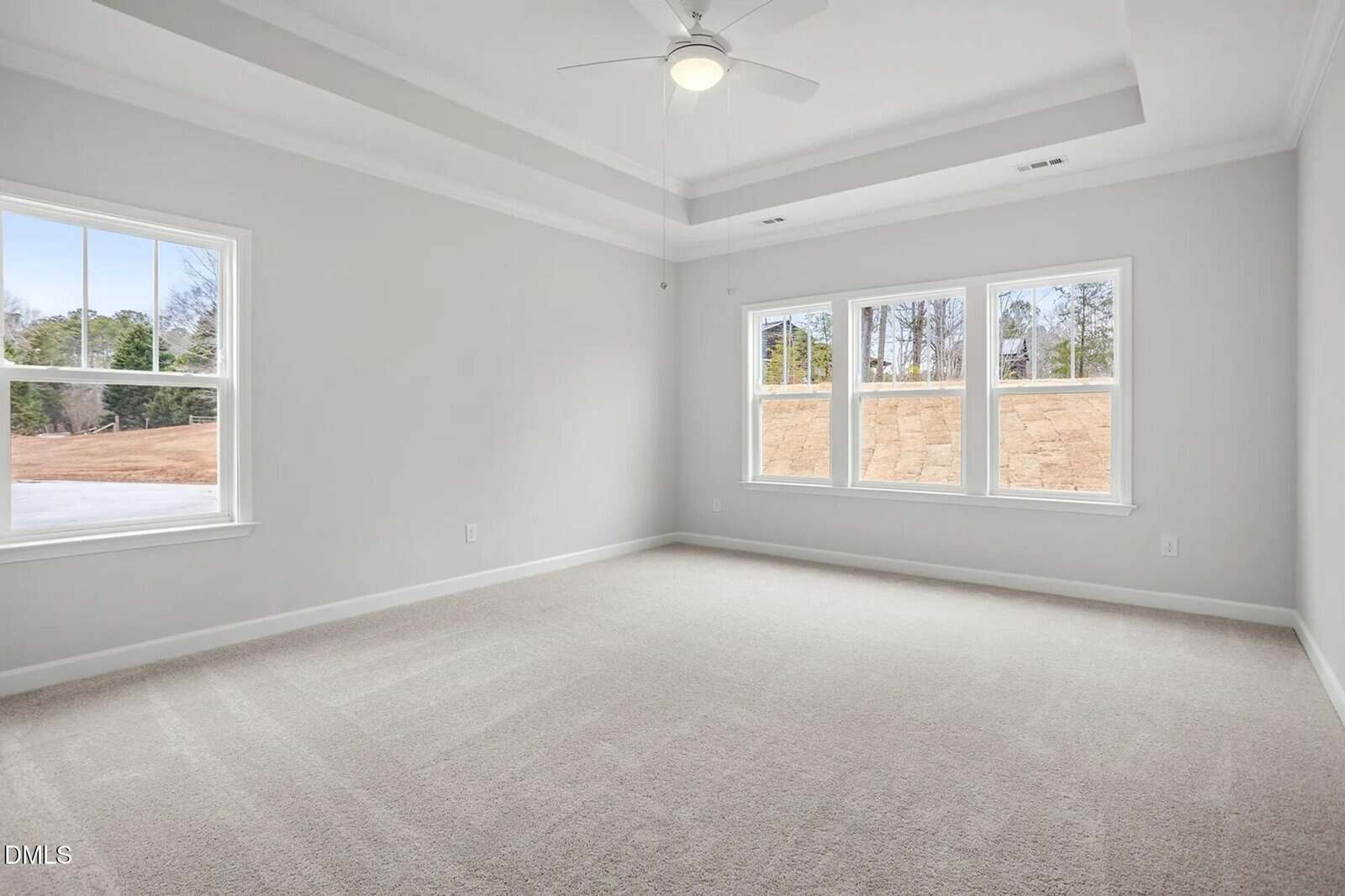 Bright, empty bedroom with beige carpet, white walls, tray ceiling, and large windows showing outdoor trees and a stone retaining wall. Ceiling fan mounted in center of the ceiling.
