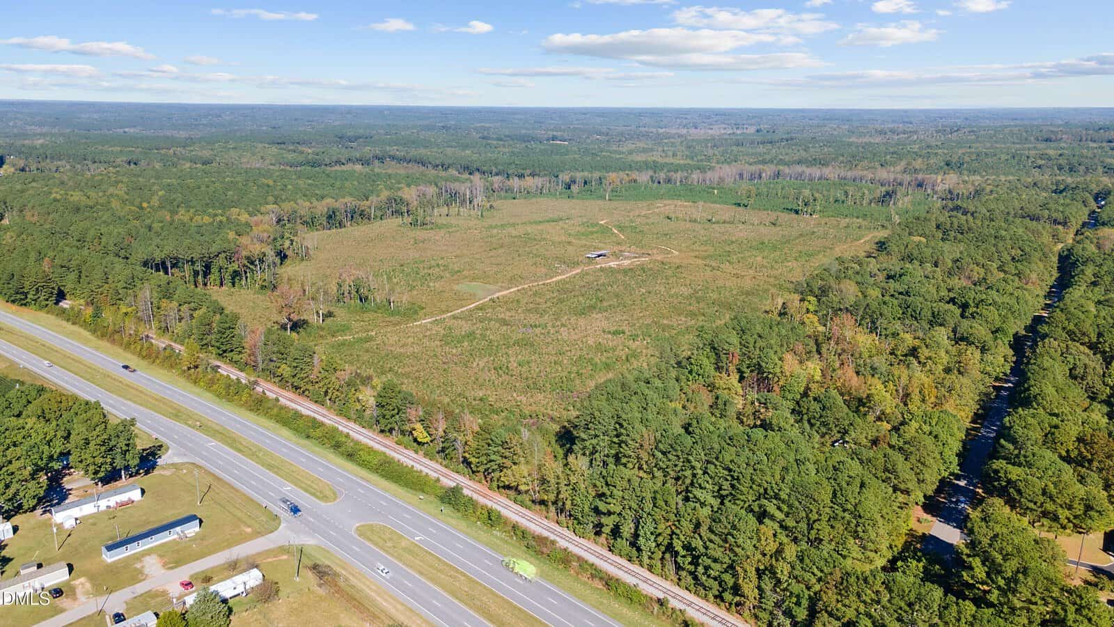 Aerial view of a large, cleared rectangular field bordered by dense forest, with a highway and mobile homes in the foreground. A small structure and a dirt path are visible within the field under a partly cloudy sky.
