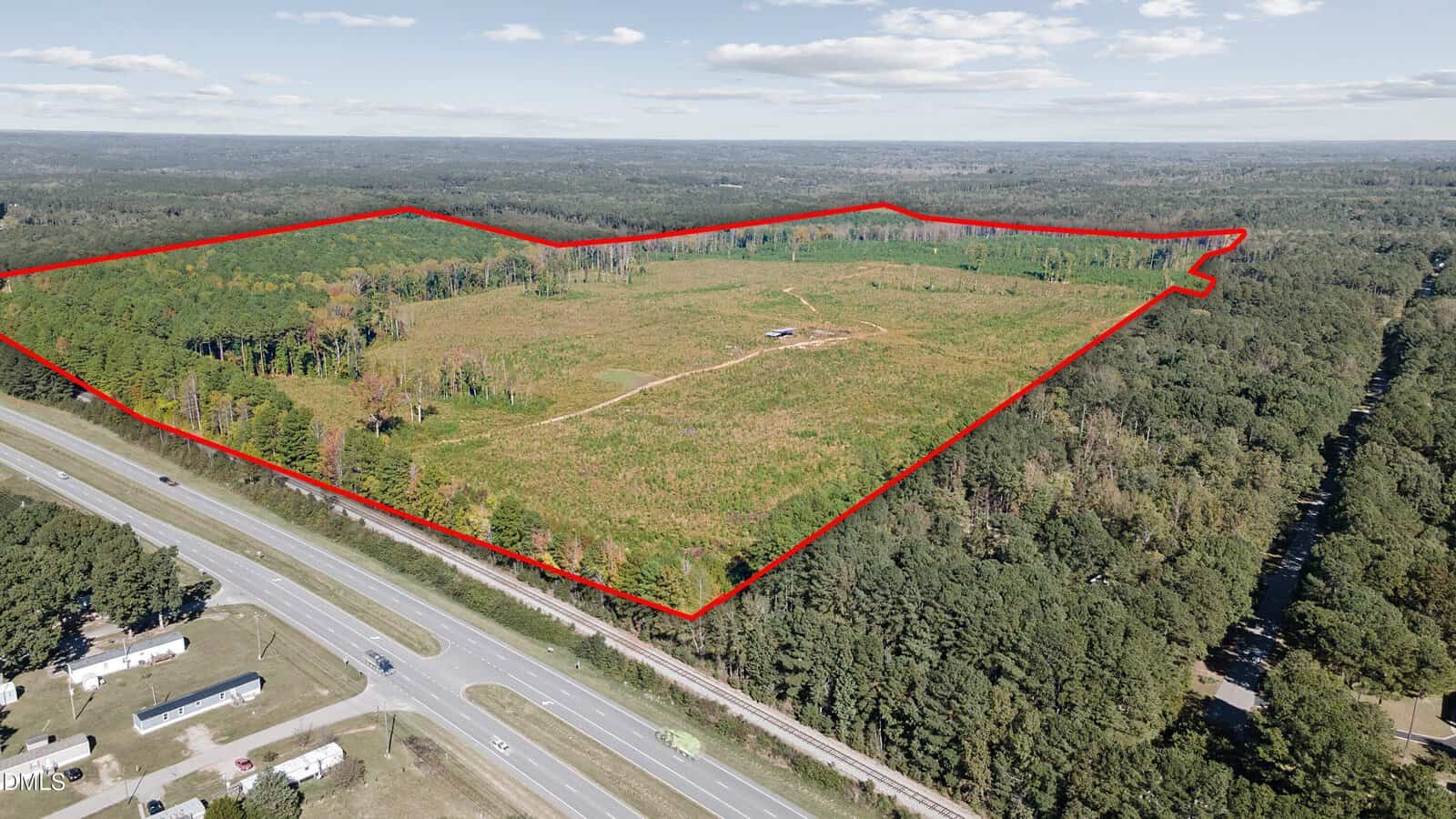 Aerial view of a large, partially cleared land area outlined in red, surrounded by dense forest. A road with vehicles runs along the foreground, and a small structure is visible near the center of the outlined area.