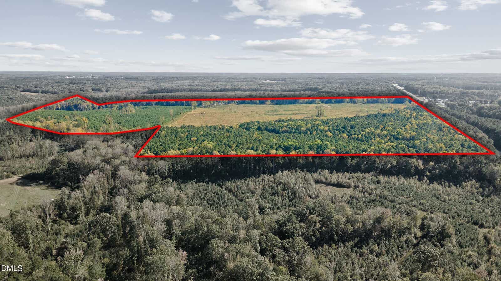 A large aerial view of a wooded and partially cleared landscape, with a section outlined in red to indicate a specific area of land. The sky is mostly clear with scattered clouds.