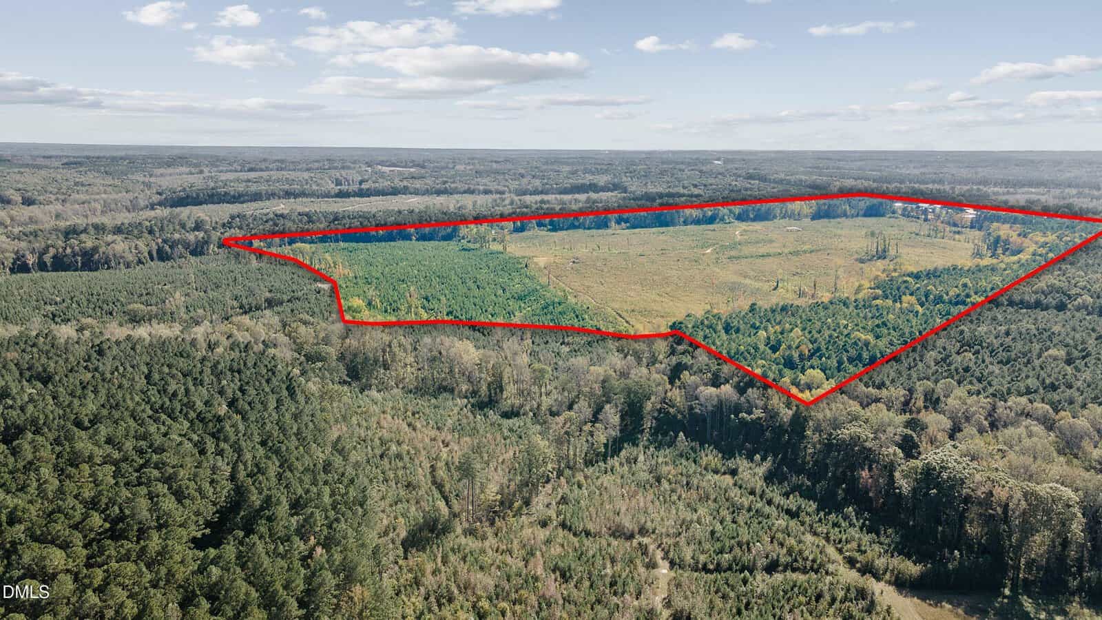 Aerial view of a large, irregularly shaped plot of land outlined in red, showing a mix of cleared and forested areas surrounded by dense woods under a partly cloudy sky.
