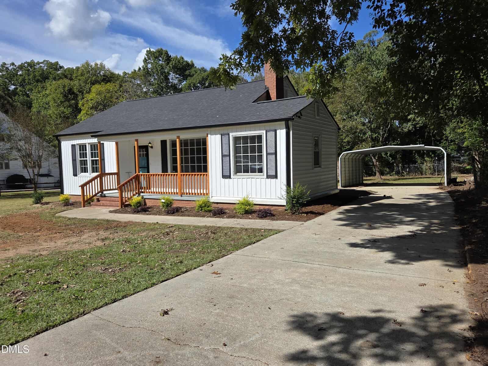 A small white house with black shutters, a front porch with wooden railings, and a concrete driveway leading to a covered carport. The yard is partially grassed with some landscaping near the house.