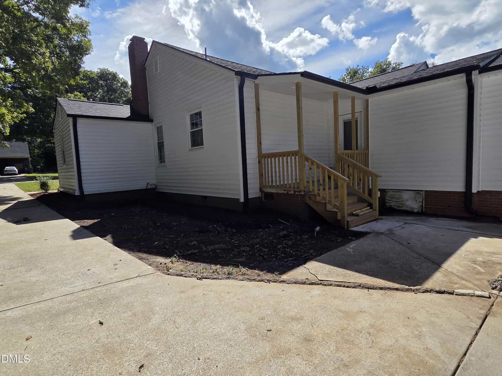 A white single-story house with a new wooden porch and steps leading to a door. The yard area near the porch has bare dirt, and the driveway is concrete. Trees and a blue sky with clouds are in the background.