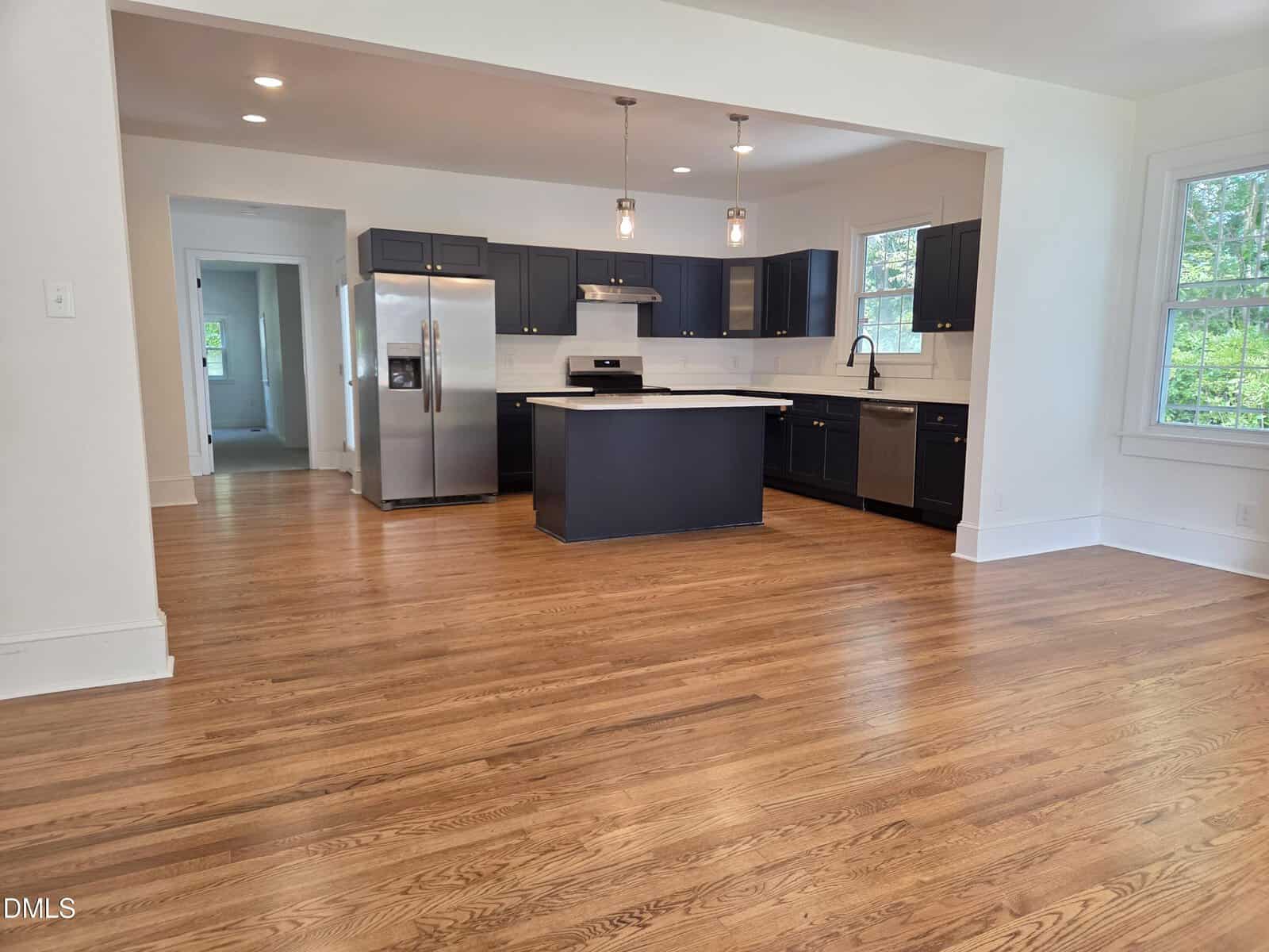 Open kitchen with dark cabinets, stainless steel appliances, center island, and white tile backsplash. Kitchen opens to a large room with hardwood floors. Multiple windows let in natural light.