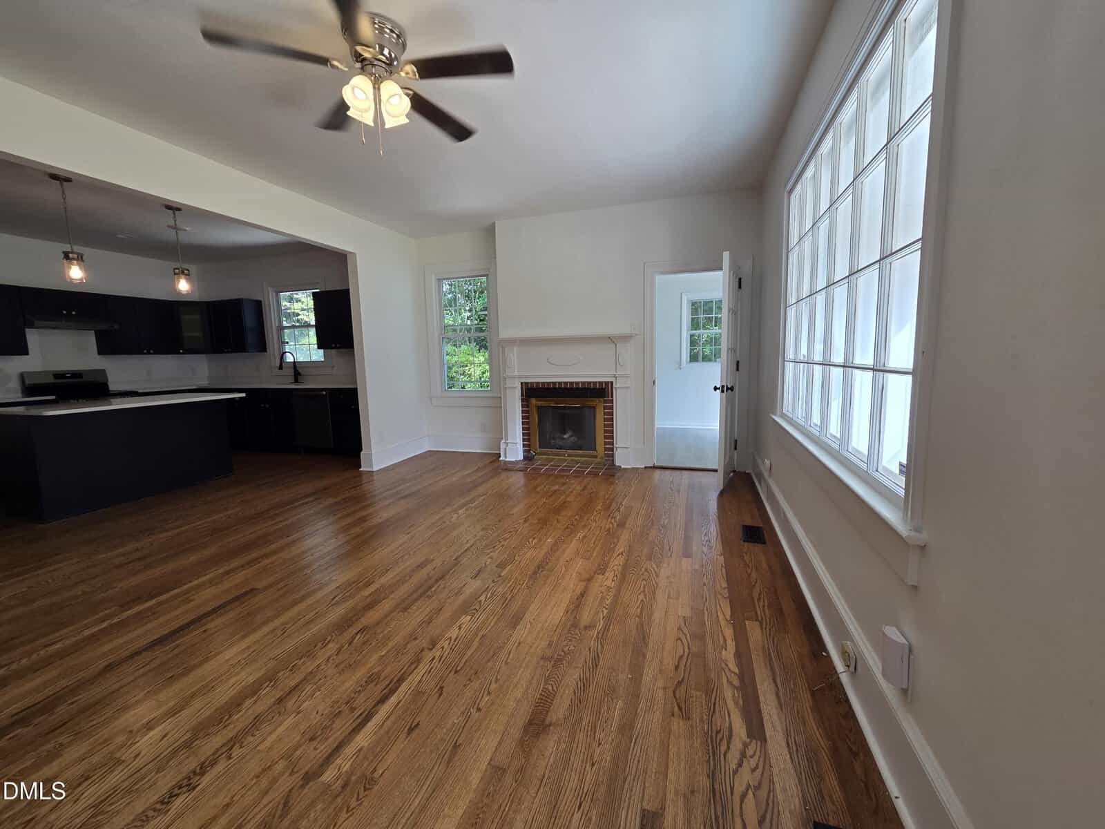 A bright living room with hardwood floors, a ceiling fan, a fireplace with a mirror above it, large windows on one wall, and an open view into a modern kitchen with dark cabinets and pendant lights.