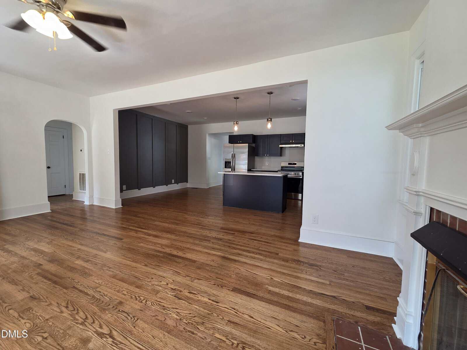 Open living space with hardwood floors, white walls, a ceiling fan, a fireplace on the right, and a modern kitchen with dark cabinets and stainless steel appliances in the background.