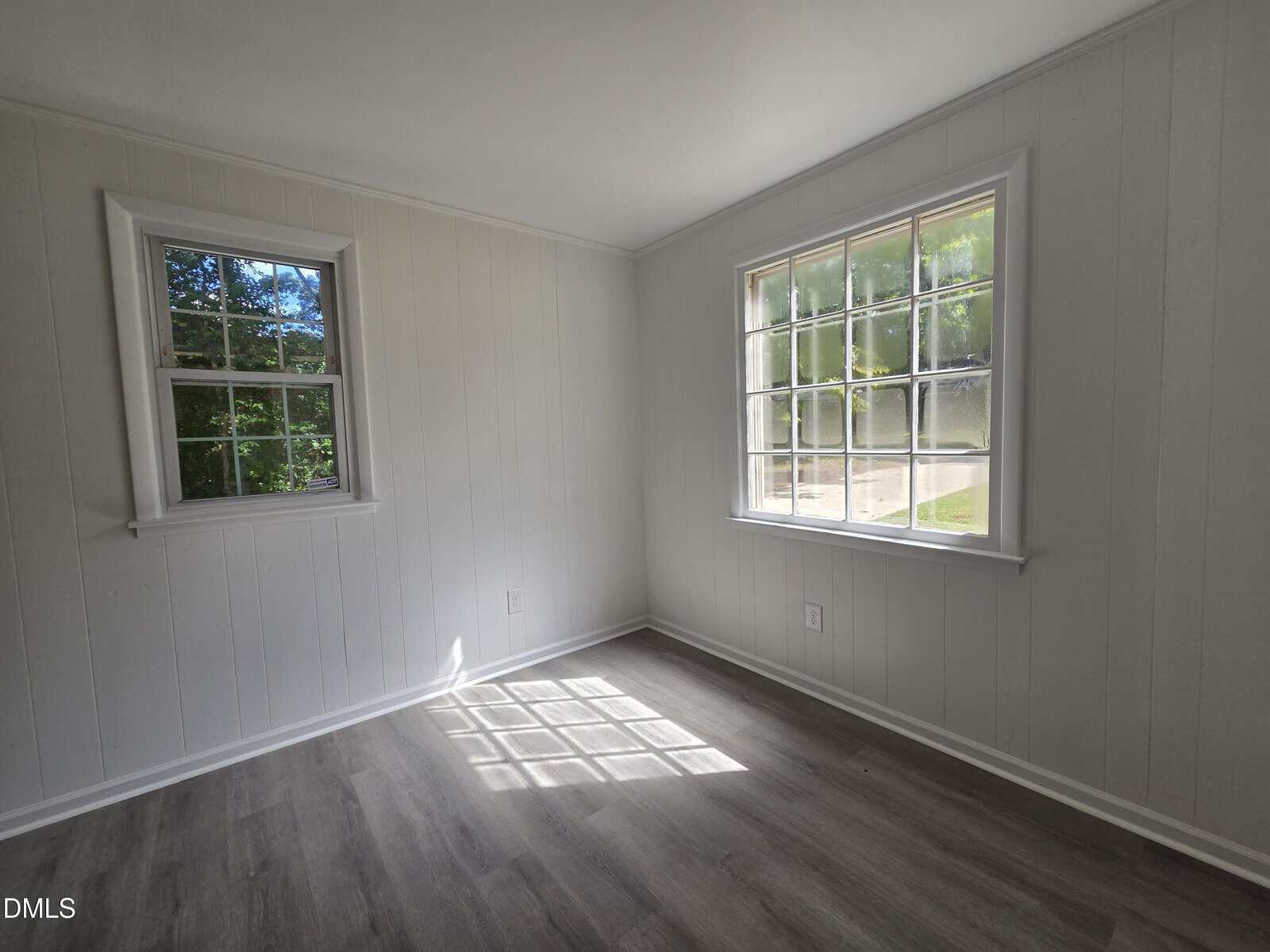 A small, empty room with light-colored wood panel walls, two windows letting in sunlight, and gray wood-look flooring. Shadows from window panes are visible on the floor.
