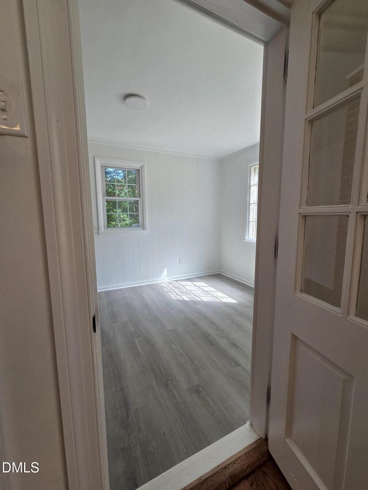 A small, empty room with light gray wood flooring, white paneled walls, two windows, and sunlight streaming in. Photo taken through a doorway with a glass-paneled door.