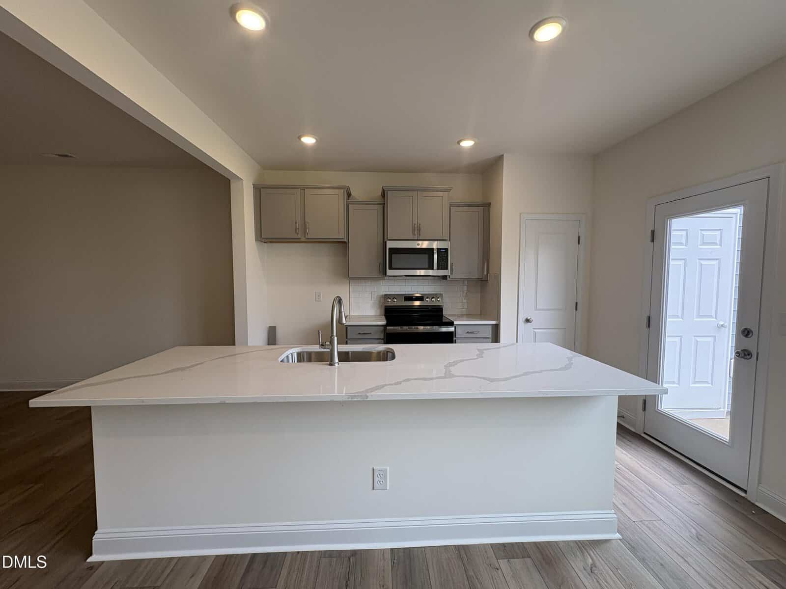 Modern kitchen with white marble island, stainless steel sink, gray cabinets, and stainless steel appliances. There is a door to the right and wood flooring throughout the space.