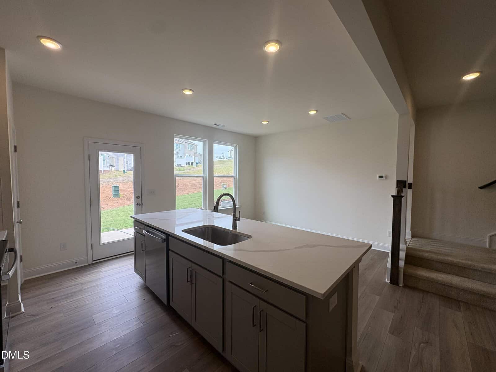 Modern kitchen with island, stainless steel appliances, light-colored countertop, and wood-look flooring. A door and windows lead to a backyard. Carpeted stairs are visible to the right.