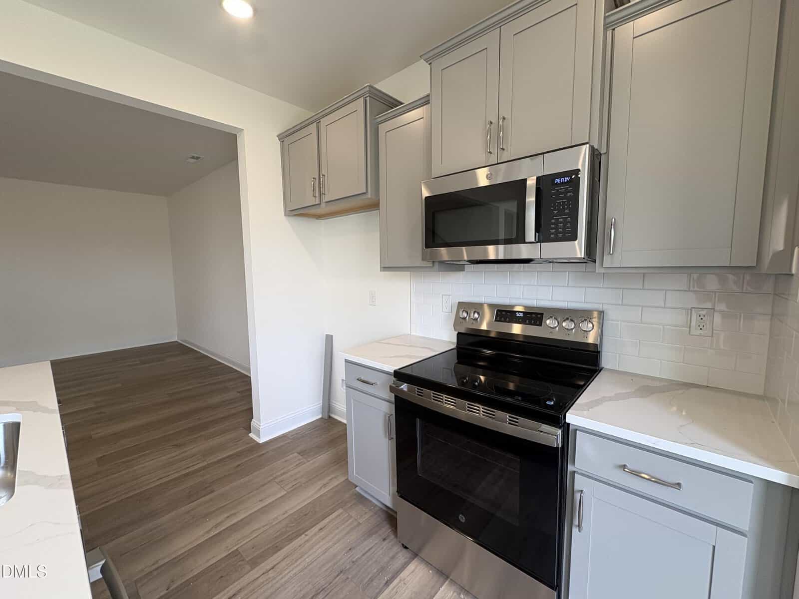 Modern kitchen with light gray cabinets, stainless steel stove and microwave, white subway tile backsplash, and wood-style flooring. An open doorway leads to another room with the same flooring.