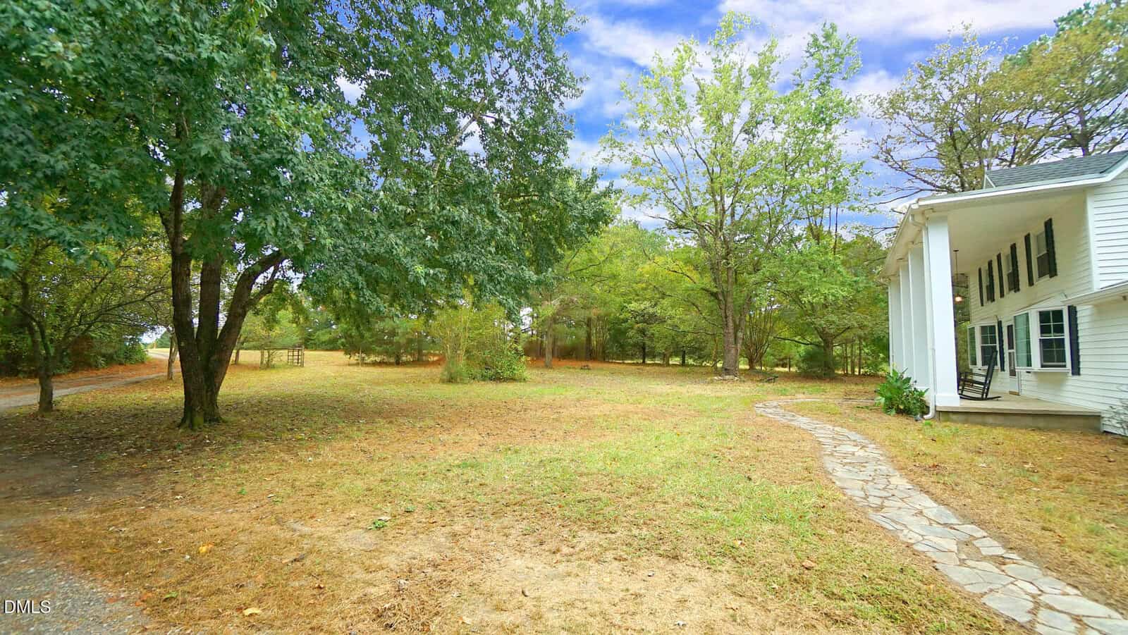 A stone walkway curves through a dry, grassy yard toward a white house with columns and a porch. Trees border the lawn and the sky is partly cloudy.