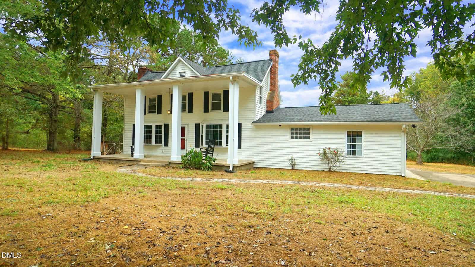 A two-story white house with tall columns, black shutters, and a brick chimney sits on a grassy lot surrounded by trees under a partly cloudy sky at 470 Flat Rock Church Road in Louisburg. A curved walkway leads to the front porch.