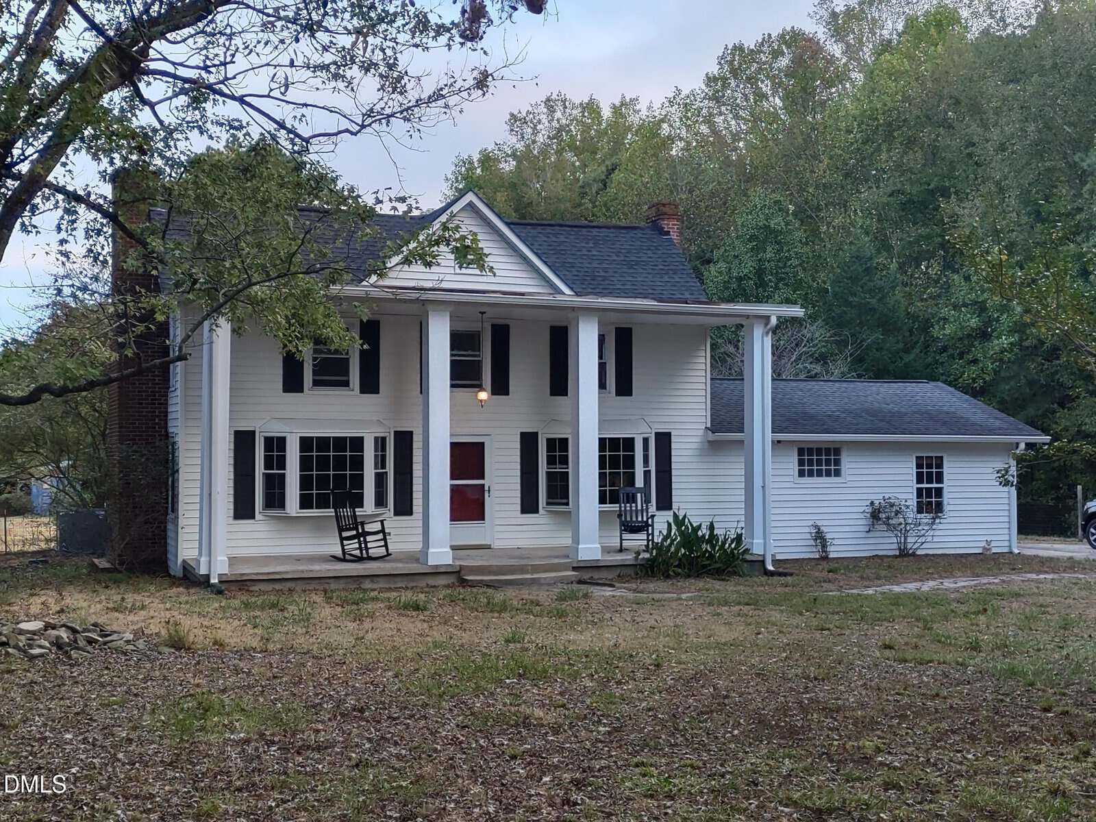 Two-story white house with large columns, black shutters, and a red front door. There are two rocking chairs on the front porch and trees surrounding the house. The yard is covered with dry grass and leaves.