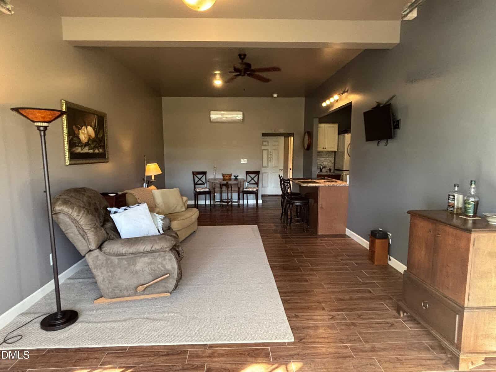 A living space with a recliner, rug, and floor lamp on the left, a dining table with four chairs in the middle, and a kitchen with barstools on the right. The room has wood flooring and neutral-colored walls.