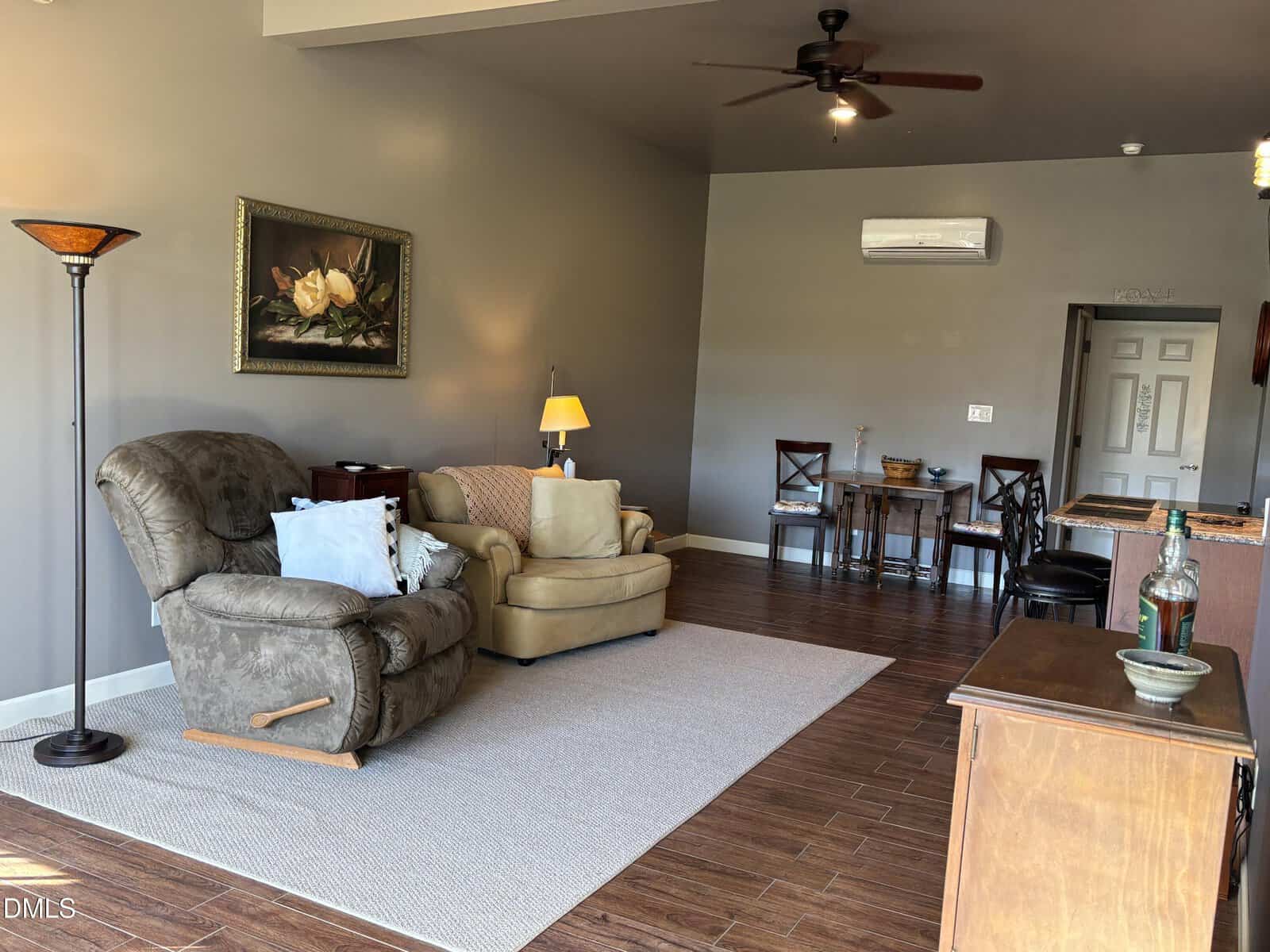 A living room with a recliner, an armchair, a standing lamp, a painting on the wall, a small dining table with chairs, wood-style flooring, and neutral-colored walls. A ceiling fan and air conditioner are visible.
