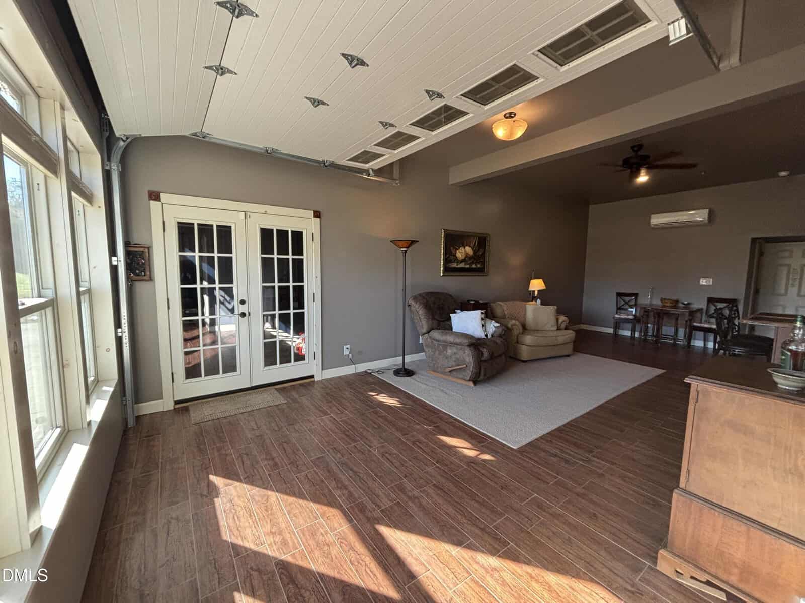 A spacious living area with wood-look tile flooring, a sectional sofa, recliner, floor lamp, and French doors. The room has large windows, a ceiling fan, and a garage-style ceiling with exposed tracks.