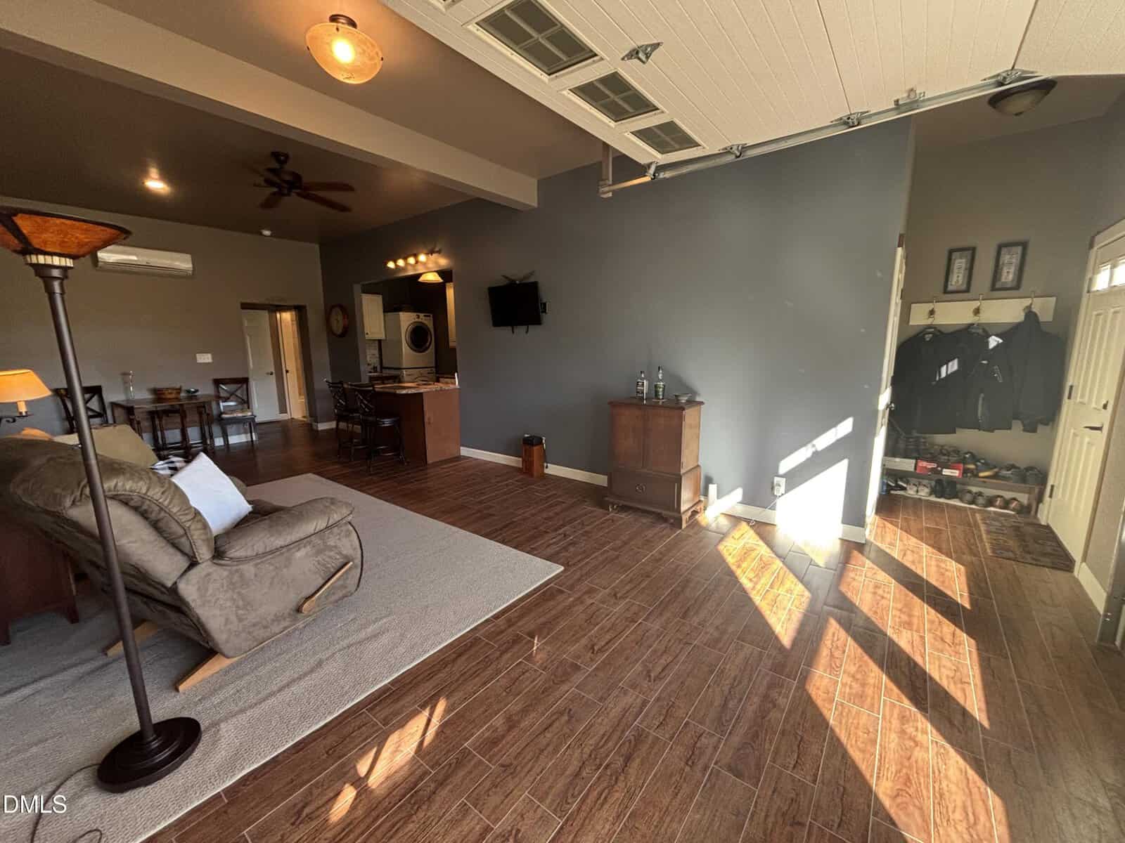 A living space with wood-look tile flooring, gray walls, and a partially open garage door. The room includes a recliner, dining area, wall-mounted TV, bar cabinet, coat hooks, and a window with sunlight streaming in.