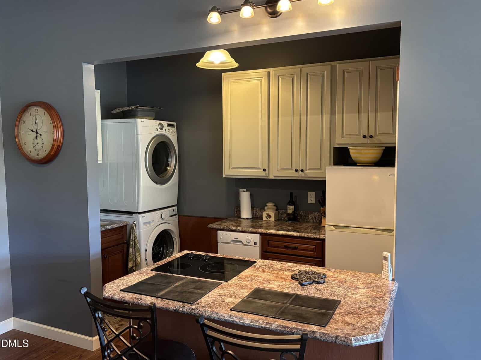 A small kitchen with a marble-patterned countertop, two chairs, a stacked washer and dryer, white cabinets, a refrigerator, dishwasher, stovetop, and a wall clock. There are lights above the counter.