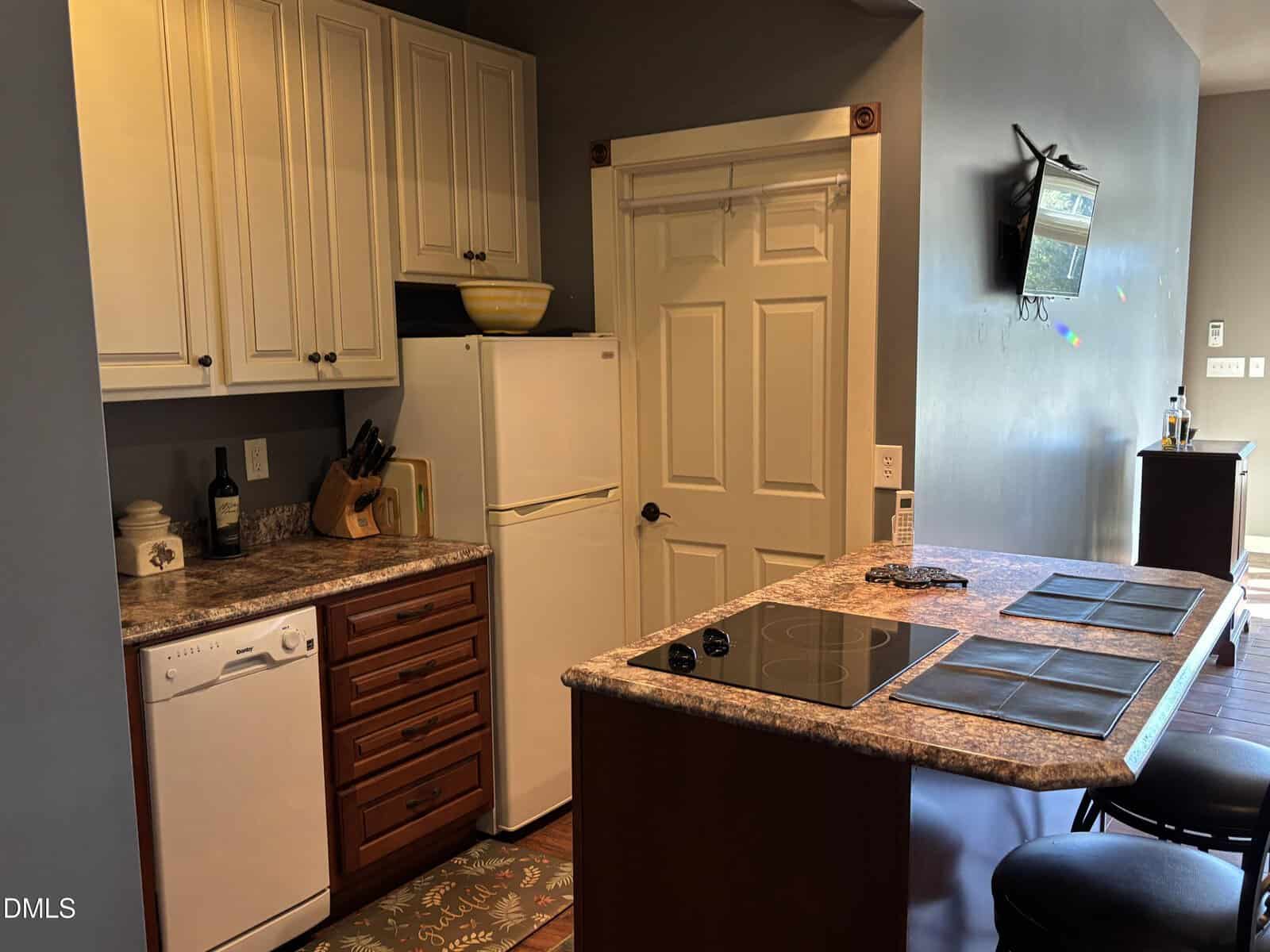 A small kitchen with white upper cabinets, dark lower drawers, a white refrigerator, a dishwasher, a countertop with an electric cooktop, and an island with stools. A wall-mounted TV is visible on the adjoining wall.