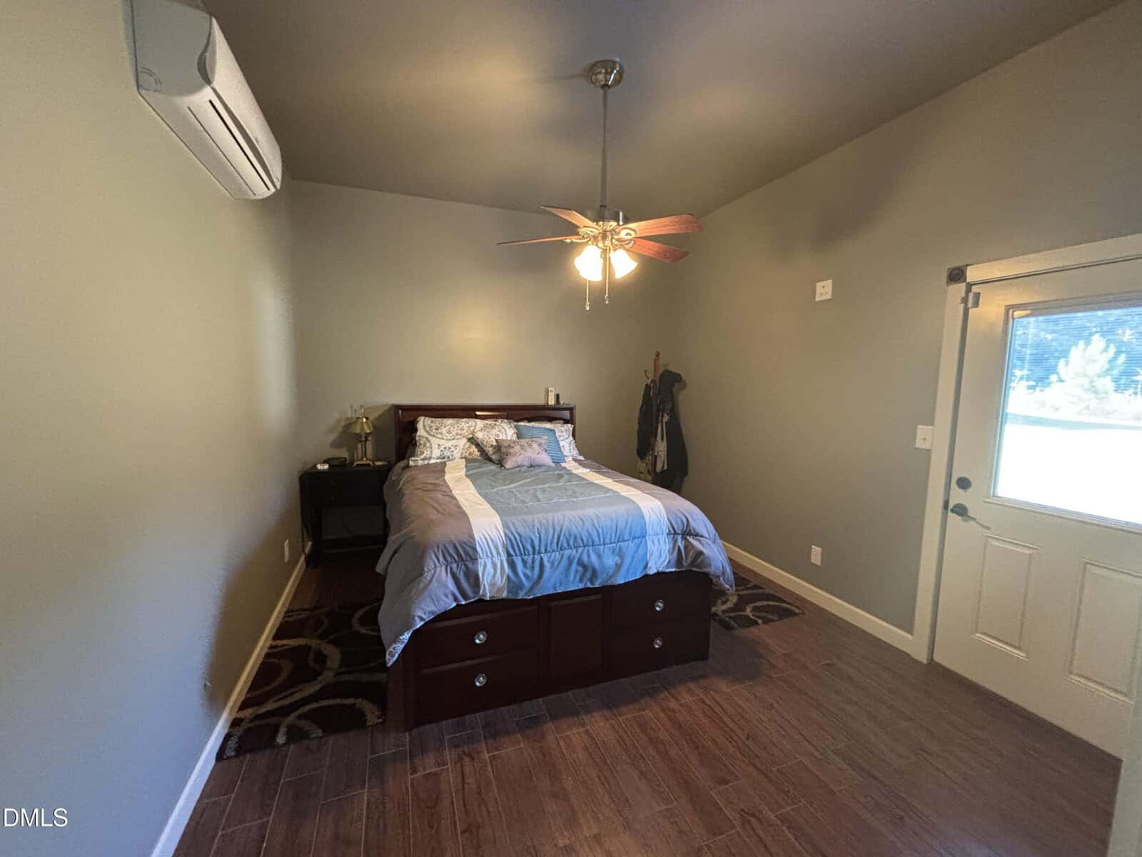 A small bedroom with a bed, nightstand, ceiling fan, and coat rack. There is a door with a window, an air conditioning unit on the wall, wood flooring, and neutral-colored walls.