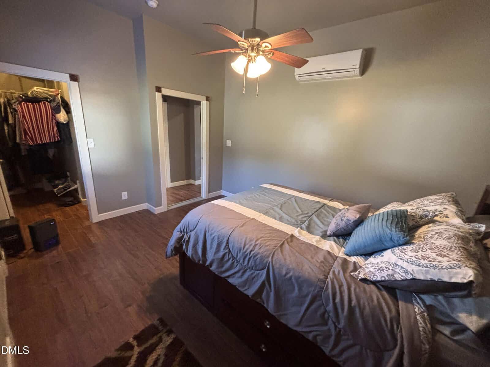 A bedroom with wood-look floors, grey walls, and a bed with blue and grey bedding. A ceiling fan, wall-mounted air conditioning unit, and two doorways—one leading to a walk-in closet and another to a hallway—are visible.