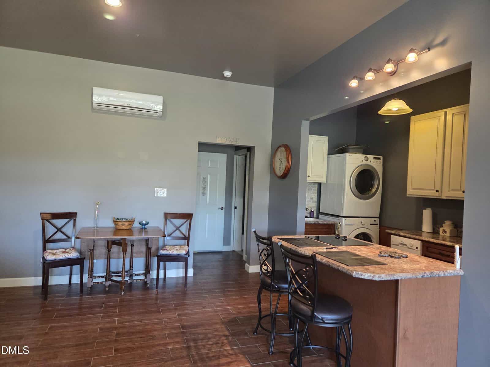 A kitchen and dining area with dark wood floors, a small dining table with three chairs, a bar with three stools, and a stacked washer and dryer in a nook next to white cabinets.