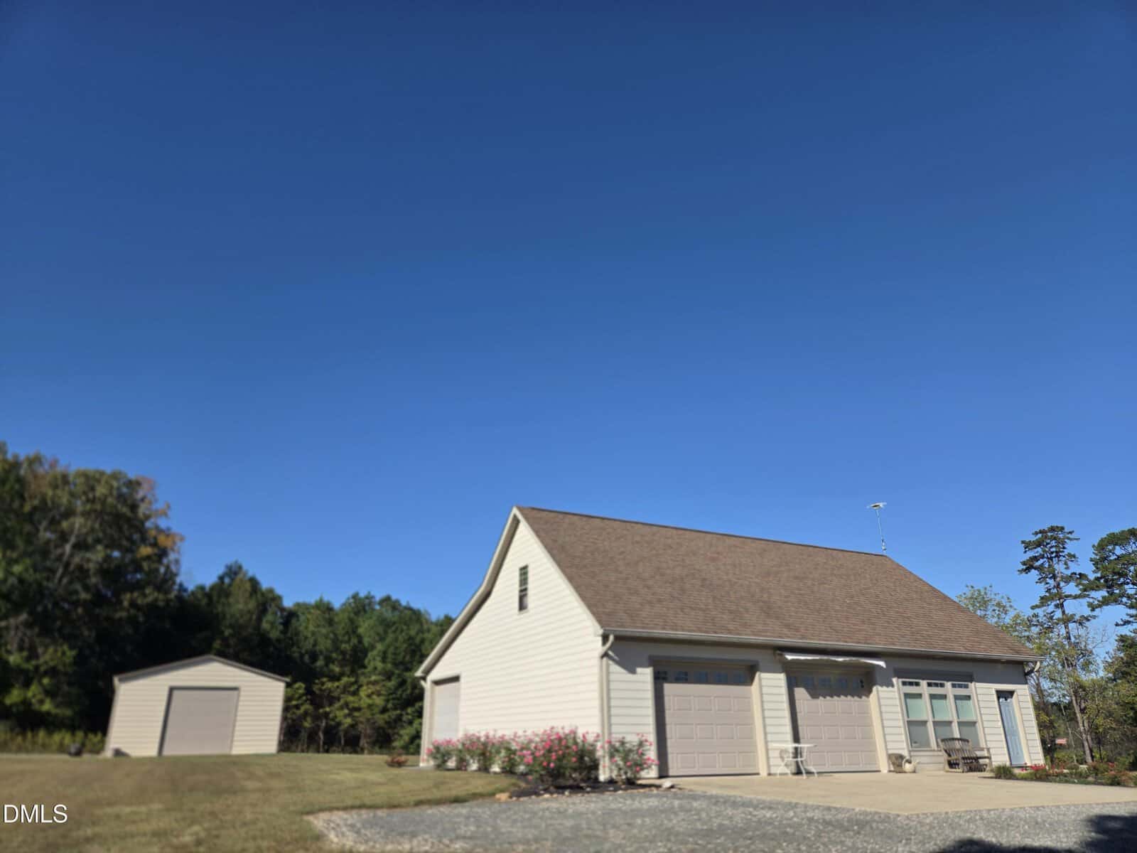 A beige detached garage with three large doors and brown roof sits on a gravel driveway, surrounded by a grassy yard with a small shed in the background and trees under a clear blue sky.