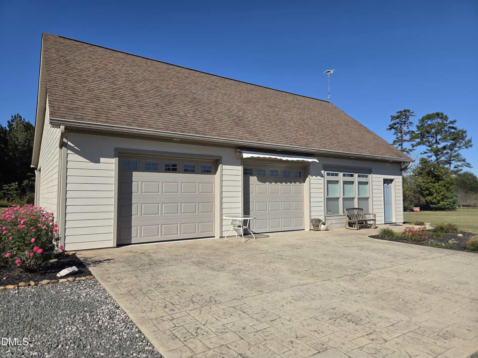 A light-colored detached garage with two closed doors, three windows, outdoor seating, and a paved driveway. There are bushes with pink flowers on one side and open grassy space in the background under a clear blue sky.