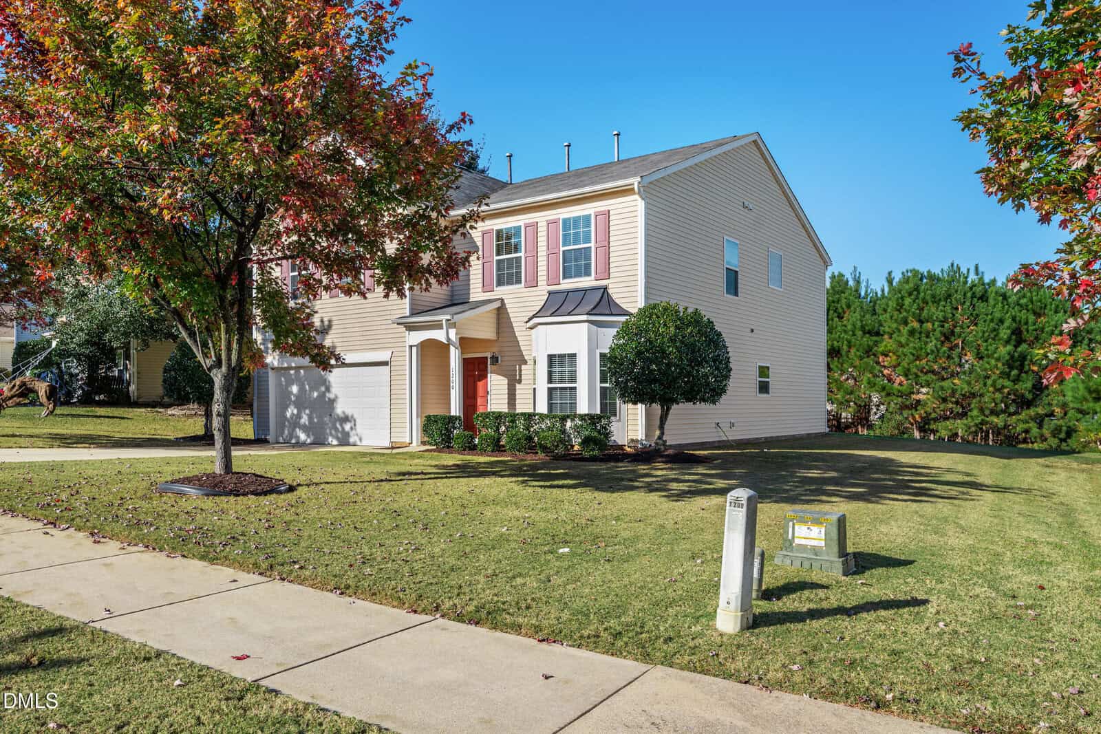 A two-story beige house with white trim and a red front door sits at 1200 Crendall Way, Wake Forest, on a neatly maintained lawn with a sidewalk, tree, and green utility boxes in front. There are trees and bushes along the side and in the background.