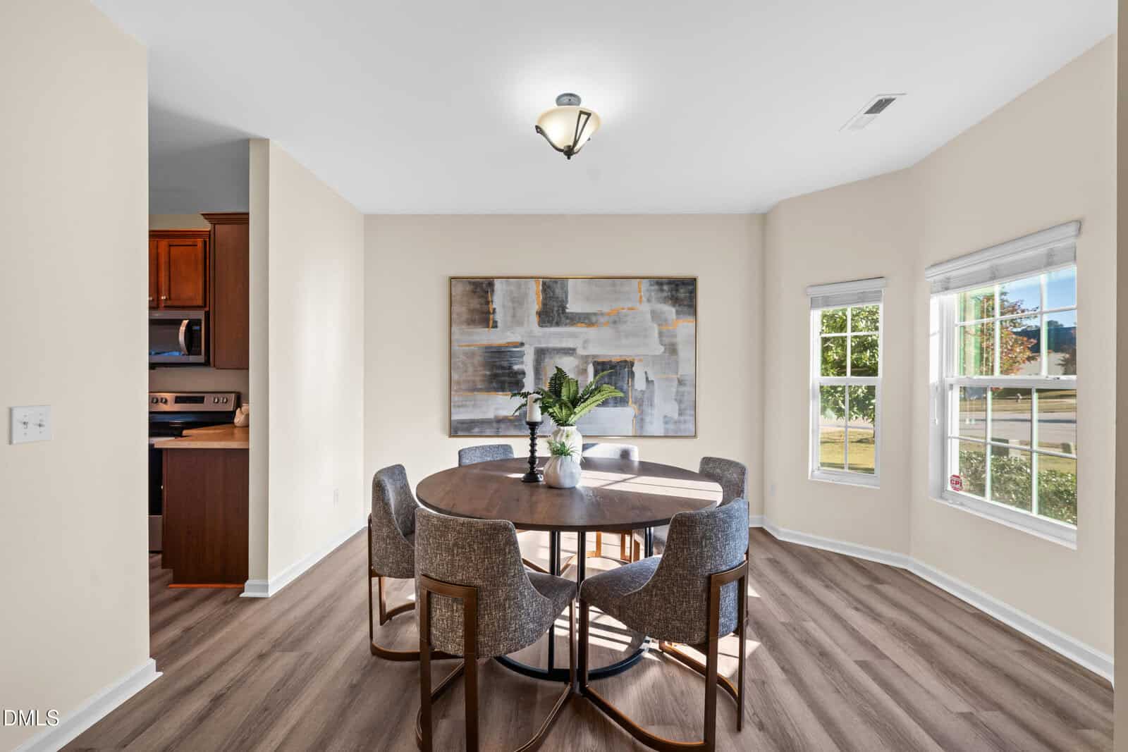 A dining area with a round wooden table, six gray upholstered chairs, a large abstract painting on the wall, and windows letting in natural light. The space has light walls and wood-style flooring.