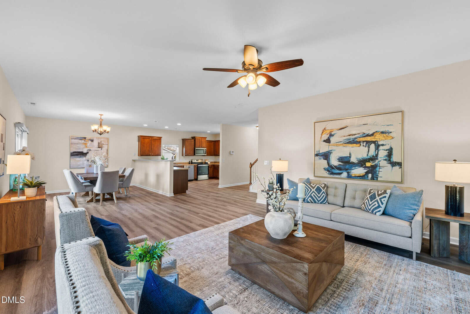Spacious open-concept living and dining area with wood flooring, neutral walls, and a ceiling fan. The room features modern furniture, decorative plants, artwork, and a view into the adjacent kitchen.
