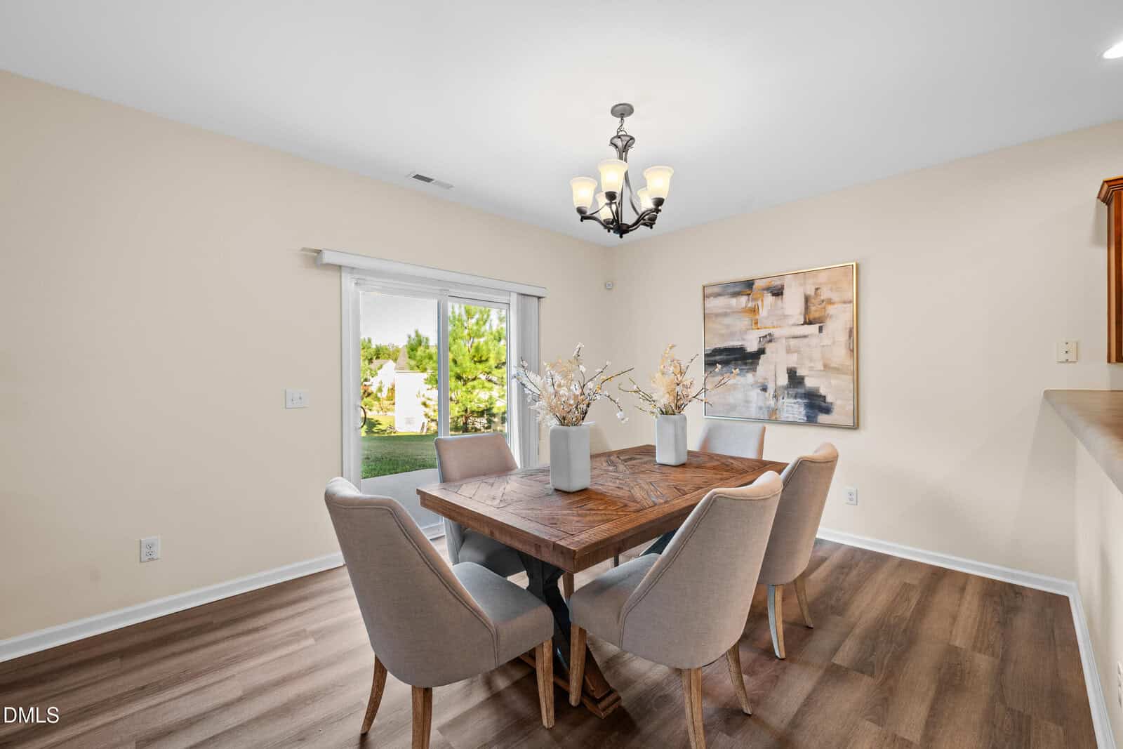 A dining room with a wooden table, six beige upholstered chairs, three white vases with decorative branches, a modern light fixture, abstract wall art, and a glass sliding door showing a view of greenery outside.