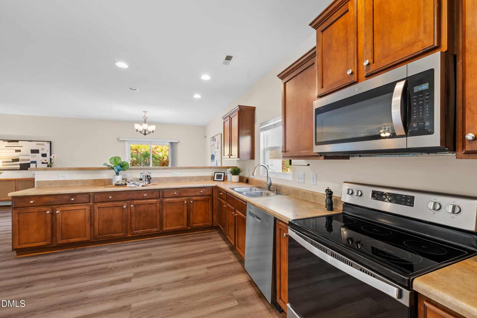 Modern kitchen with wooden cabinets, stainless steel appliances including a microwave and electric stove, double sink, beige countertops, and wood-look flooring. A dining area with a chandelier is visible in the background.