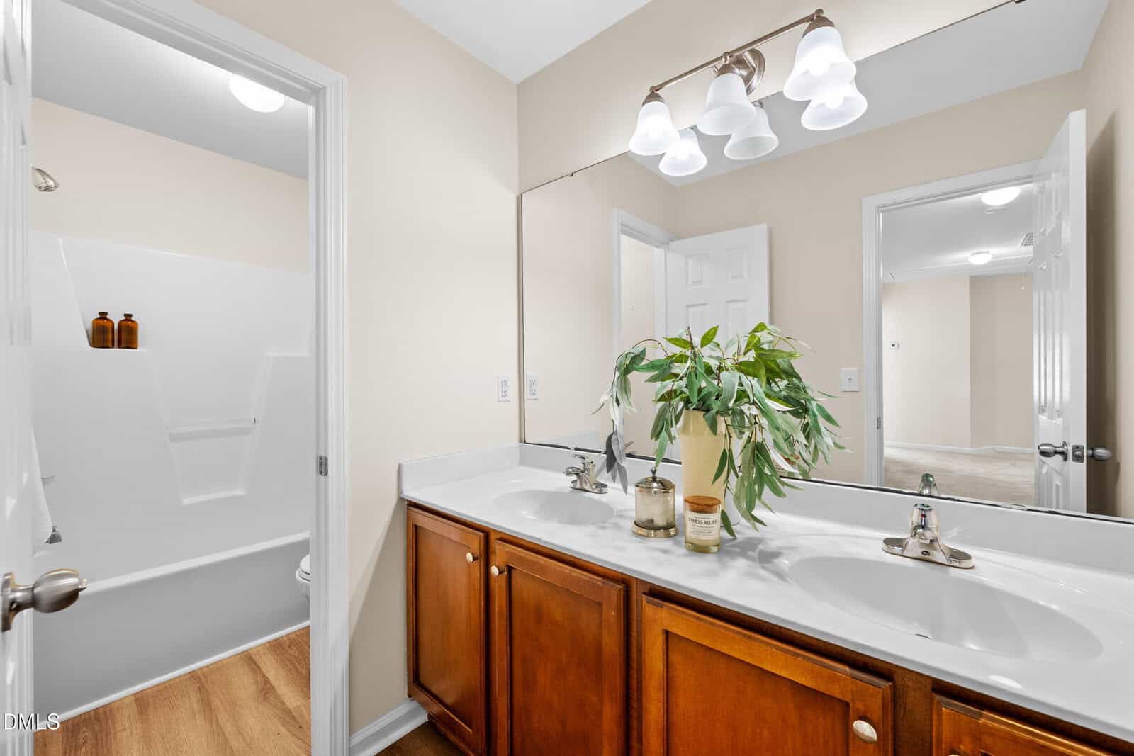 A bathroom with double sinks, a large mirror, wooden cabinets, and a plant on the countertop. A shower-tub combo is visible in the background with two brown bottles on the built-in shelf.