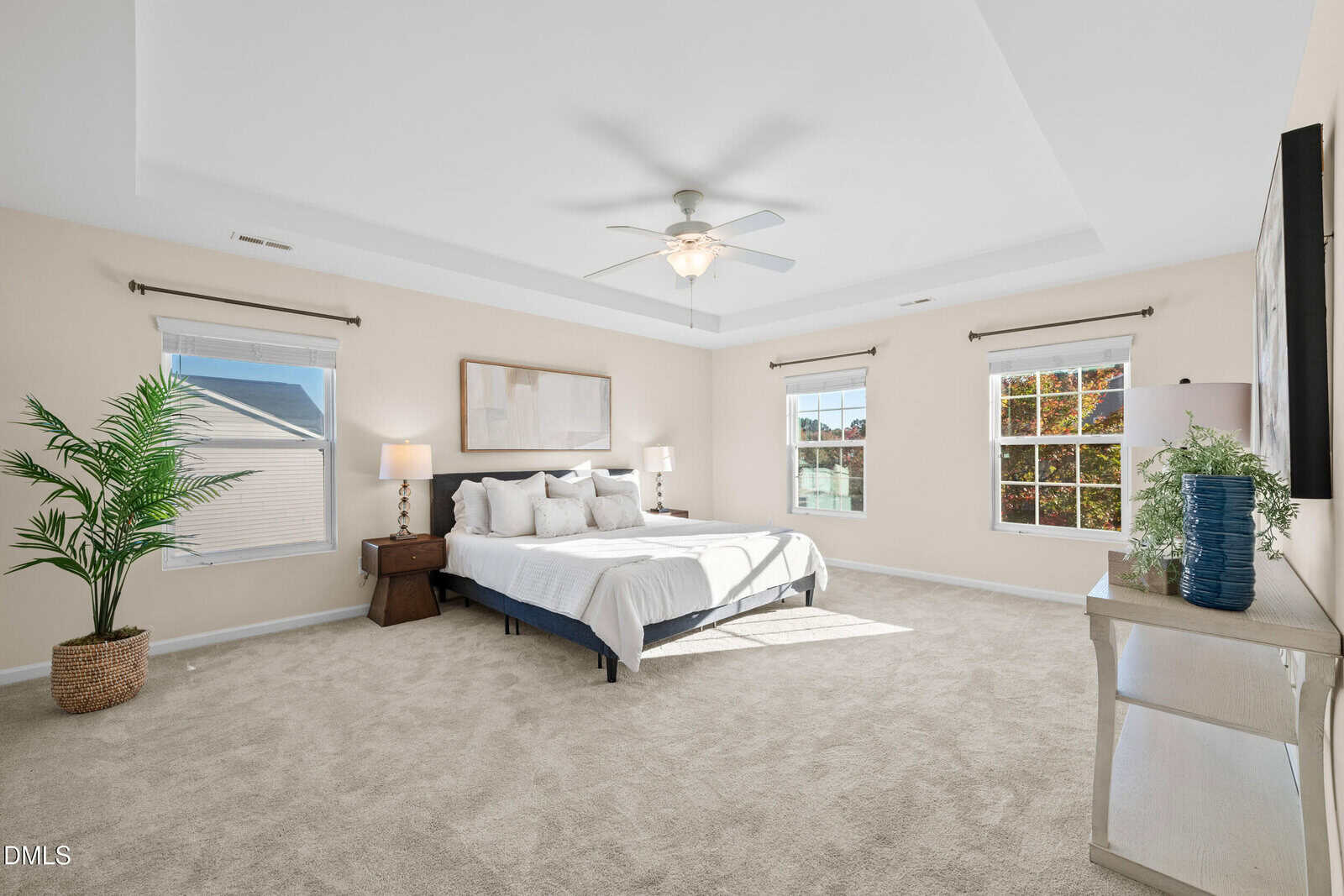 Spacious bedroom with neutral walls and carpet, a bed with white bedding and pillows, a nightstand with a lamp, large windows with white blinds, a potted plant, and a dresser with a blue vase and framed art.