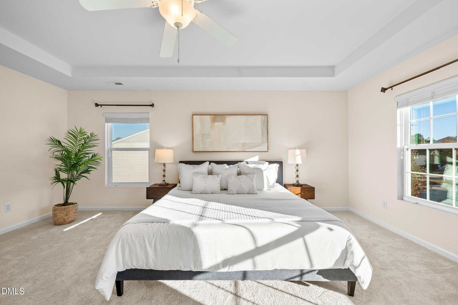 Bright bedroom with a large bed, white bedding, two side tables with lamps, a potted plant, beige walls, carpeted floor, and two windows letting in natural light.