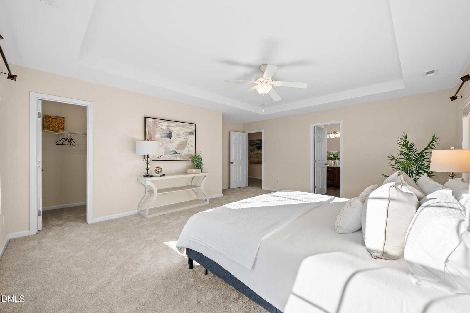 Bright bedroom with a large bed, beige walls, light carpet, ceiling fan, and natural light. Room features double doors, a closet, a console table with decor, and a potted plant in the corner.