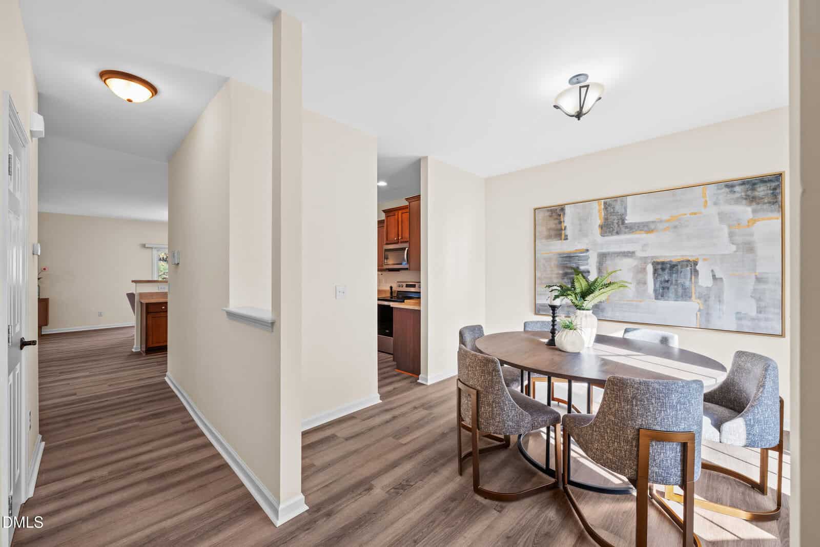 A dining area with a round wooden table, six gray upholstered chairs, a potted plant centerpiece, and a large abstract painting on the wall. The space has light beige walls and wood-style flooring.