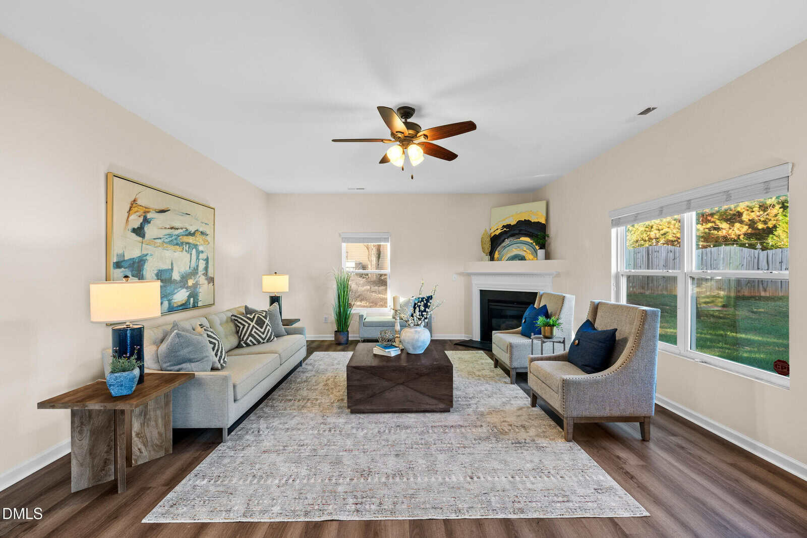 A living room with a beige sofa, two armchairs, a wooden coffee table, abstract wall art, a ceiling fan with lights, a fireplace, large rug, and windows letting in natural light. The floor is wood-patterned.