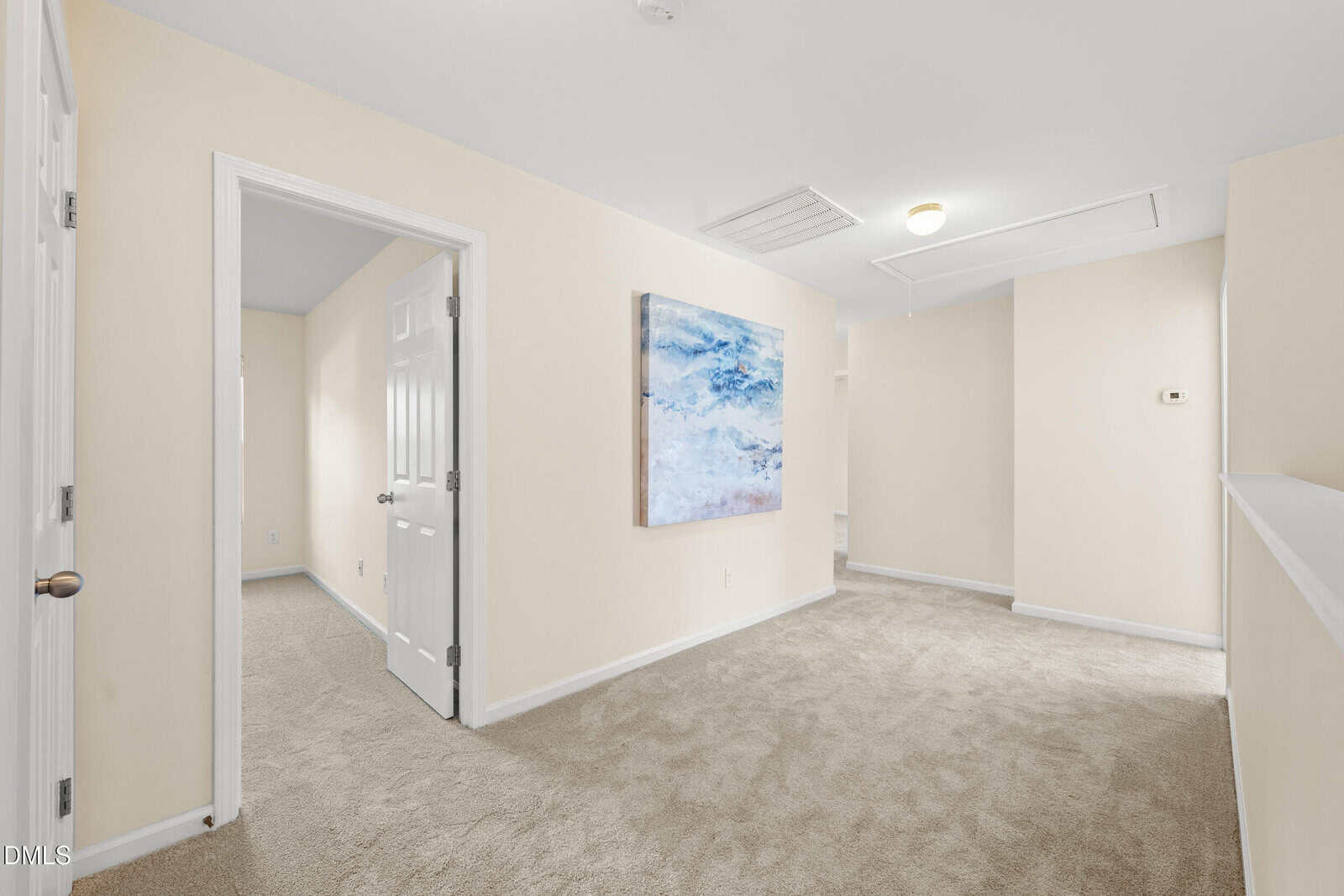 A carpeted hallway with light beige walls, a white ceiling, and a partially open door leading to another room. A large abstract painting hangs on one wall, and ceiling lights illuminate the space.